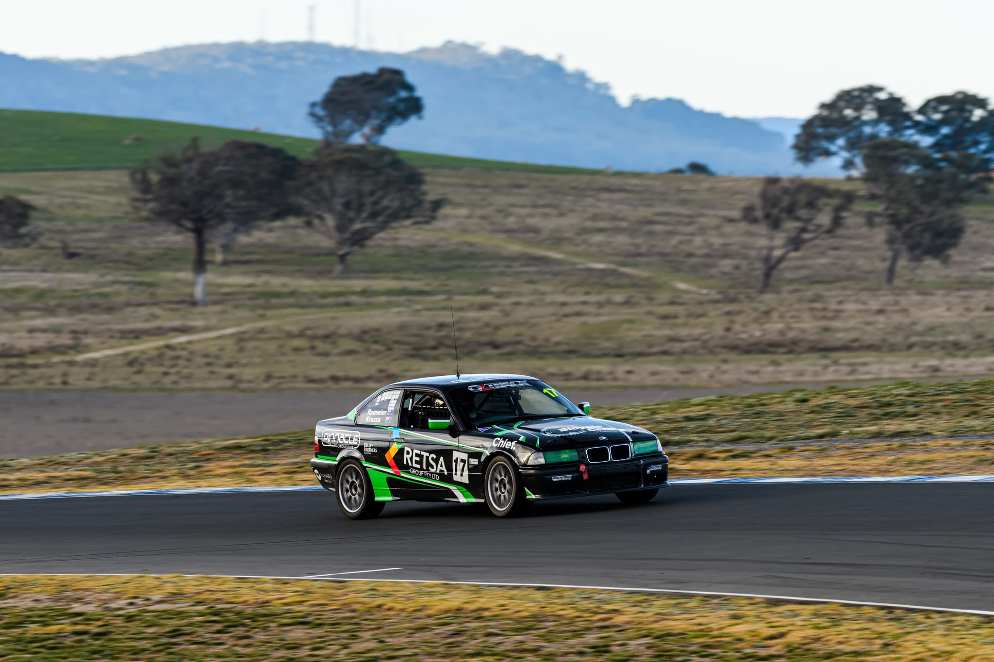 The #17 Project E36 BMW at speed on track with the Wakefield Park hills and gum trees behind during MRA Round 6