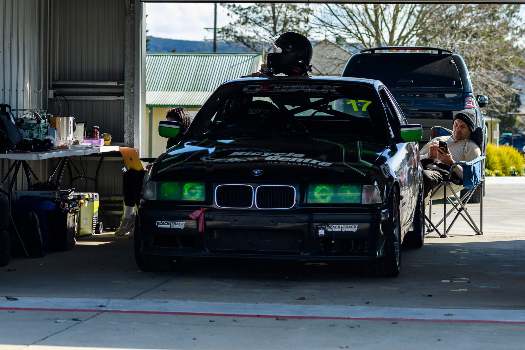 The #17 Project E36 BMW parked front-on in the garage with a helmet resting on the roof and a crew member relaxing trackside at Wakefield Park